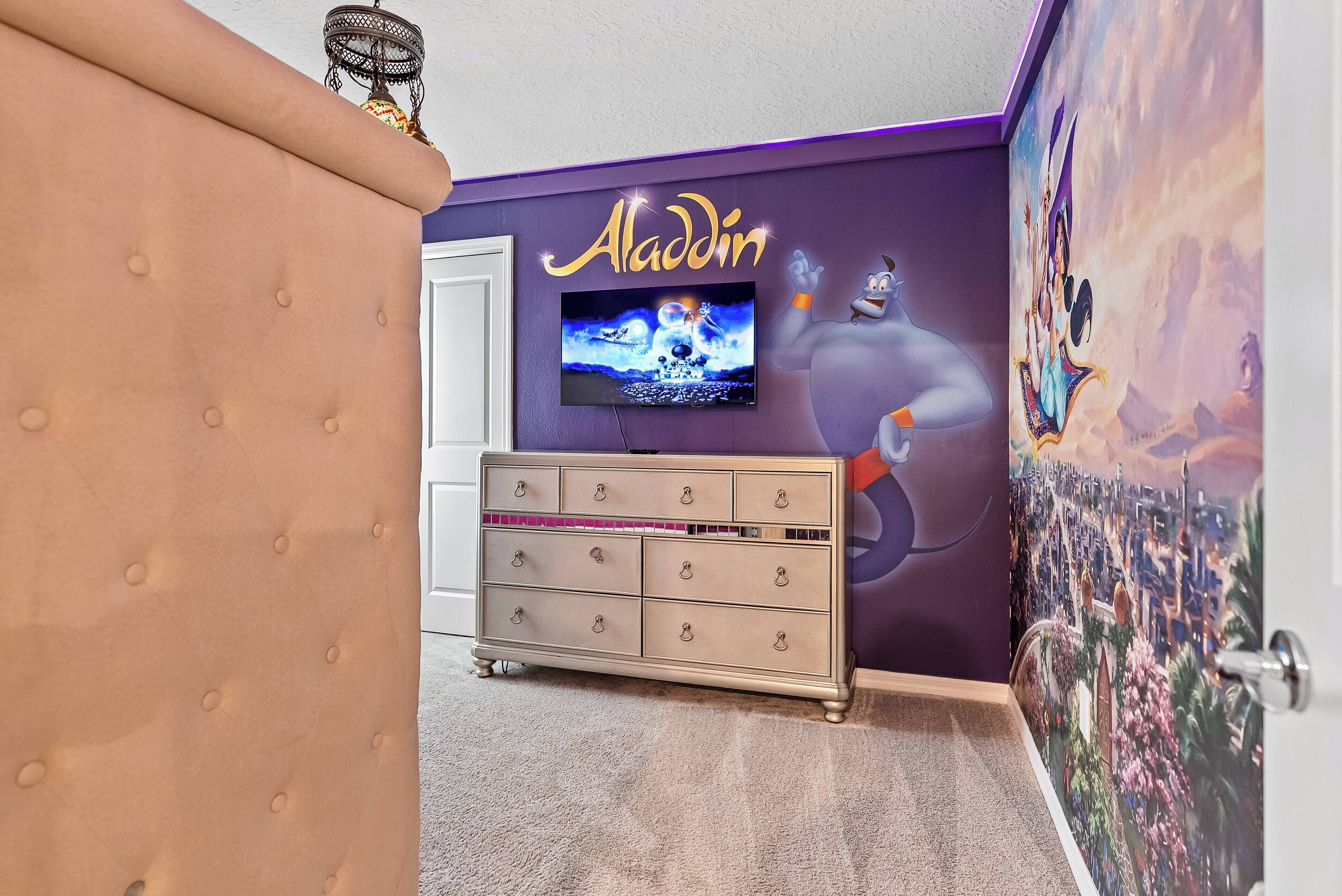 A room with a tufted headboard, a dresser with multiple drawers, a television mounted on the wall, and a mural of Aladdin's lamp.