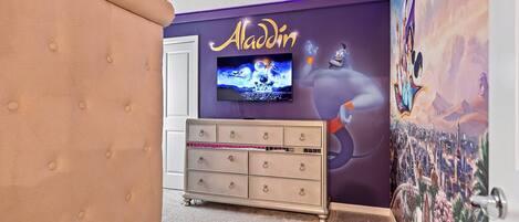 A room with a tufted headboard, a dresser with multiple drawers, a television mounted on the wall, and a mural of Aladdin's lamp.