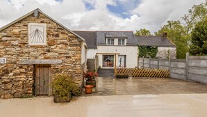 Un bâtiment en pierre avec une porte en bois et une plaque d’immatriculation, une maison blanche avec un toit gris et un espace clôturé agrémenté de plantes en pot.