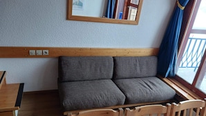 A room with a wooden bench, a grey sofa with cushions, a wooden headboard, a window with blue curtains, and a mirror on the wall. - Ski-in duplex for 6 in Arc 1800, southeast balcony (Les Arcs 1800)