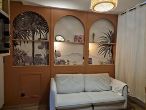 A living room with a sofa, a wall with three shelves displaying decorative items, and a hanging light fixture. - Cozy 17m² studio, near cable car, 2/4 pers, parking (Cauterets)
