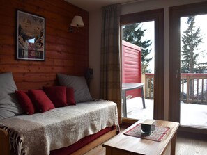 A cozy room with a wooden wall, a bed with red and grey pillows, a table with a book, and a window with a view of trees and a red building outside. - South-facing studio with parking in Font-Romeu (Font-Romeu-Odeillo-Via)