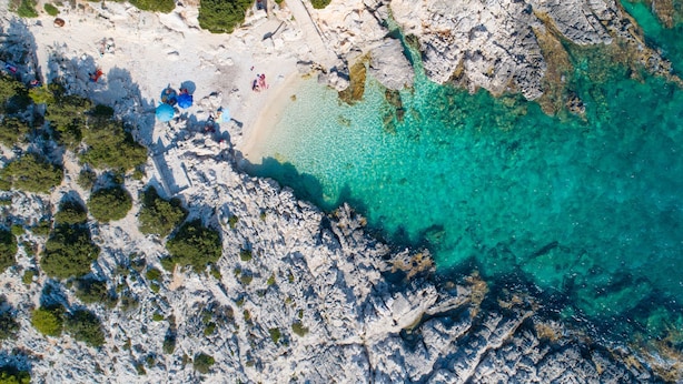Luftaufnahme einer Küstenlandschaft mit felsigem Gelände, klarem türkisblauem Wasser und spärlicher Vegetation.