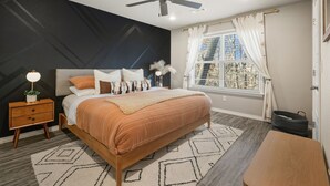Un dormitorio con cama, mesita de noche, ventilador de techo, ventana con cortinas y vistas al exterior.