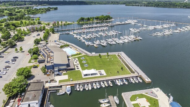 An aerial view of a marina with numerous boats docked, a parking lot, and a building with a green lawn area.