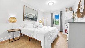 A bedroom with a bed, bedside table, lamp, artwork, dresser, and a window with curtains.