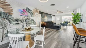 Una sala de estar moderna con una mesa de comedor de cristal, sillas blancas, un sofá oscuro y una planta grande.