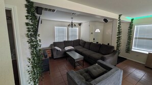A living room with a sectional sofa, a small table, and a grey armchair. There are white walls, a ceiling fan, and a window with blinds. - Candle Maker's Cove: King Bed, Fiber WiFi, Full Kitchen Access (Leander)