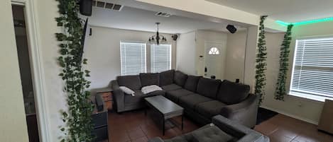 A living room with a sectional sofa, a small table, and a grey armchair. There are white walls, a ceiling fan, and a window with blinds.