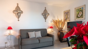 En stue med en grå sofa, to vægmonterede lamper, et fjernsyn og en stor plante.