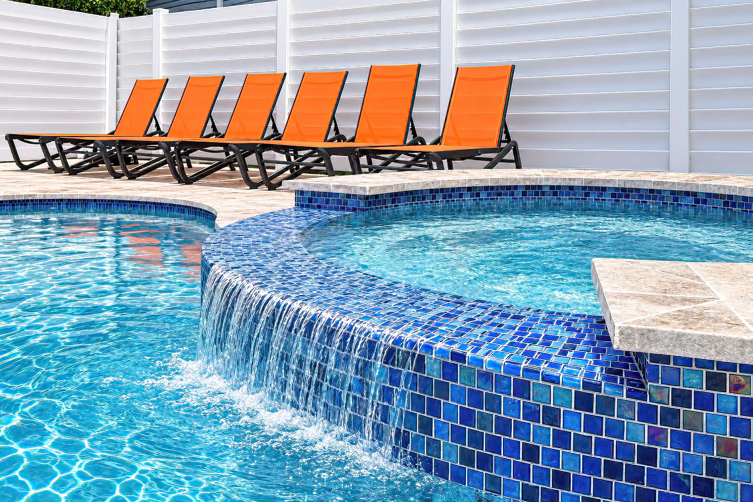 A swimming pool with a blue mosaic tile edge, surrounded by a white fence and orange lounge chairs.