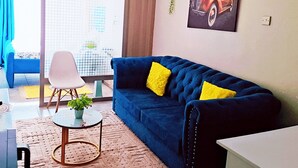 A living room with a blue sofa, a white chair, a small round table with a plant, and a large window with a view of buildings. - Affordable Studio Apartment Near JKIA, Fedha Embakasi. (Nairobi)