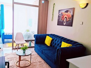 Een woonkamer met een blauwe bank, een witte stoel, een klein rond tafeltje met een plant, en een groot raam met uitzicht op gebouwen.