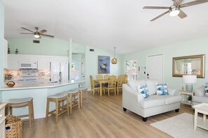 Eine moderne Küche mit weißen Geräten, ein Essbereich mit Holzsesseln und ein Wohnzimmer mit einem weißen Sofa und gemusterten Kissen.