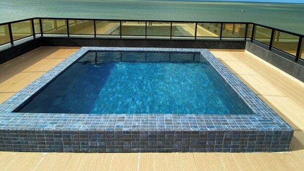 Una piscina rectangular con borde de azulejos, rodeada por un muelle de madera y una barandilla negra, con vistas a una masa de agua bajo un cielo parcialmente nublado.