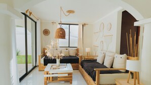 A modern living room with a sofa, a coffee table, and a side table. There is a window with curtains, a hanging light fixture, and wall art. - House with Private Pool - 3BR Cancun Downtown Zone (Cancún)