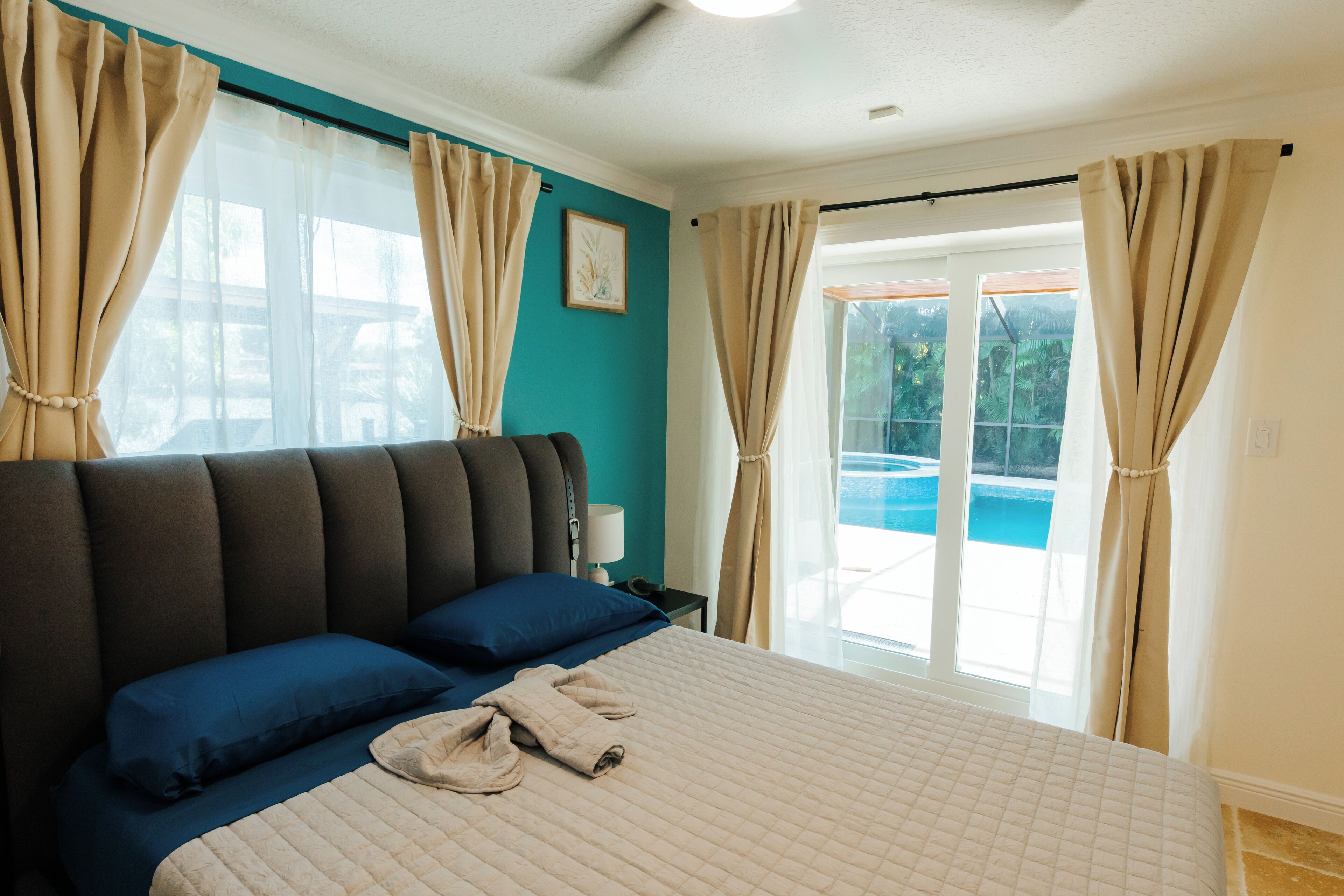Une chambre avec un grand lit, deux oreillers, une table de chevet et une vue sur une piscine, visible à travers une porte vitrée.