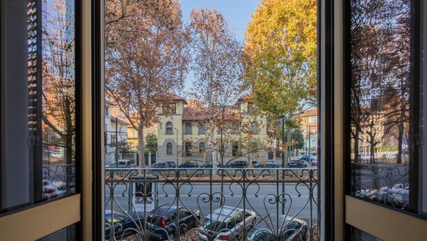 Vista attraverso una finestra con ringhiera in metallo, con auto parcheggiate e alberi dai colori autunnali.