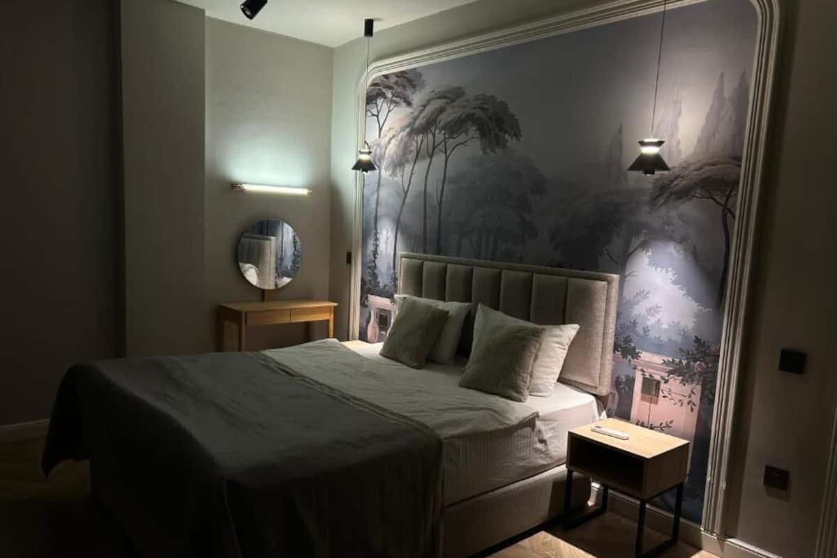 Quarto com cama, mesas de cabeceira, espelho e um mural na parede.