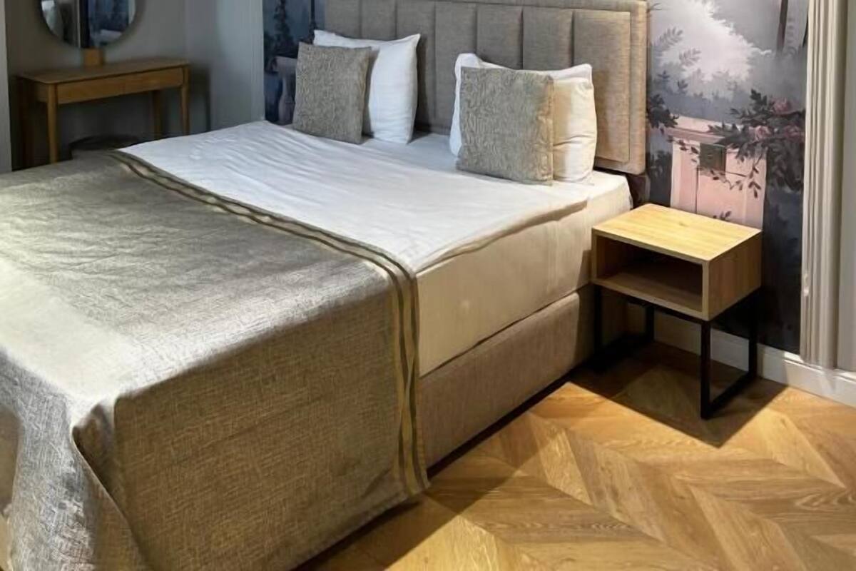Quarto com uma cama grande, uma mesa de cabeceira, uma luminária de parede e um mural com árvores e montanhas.