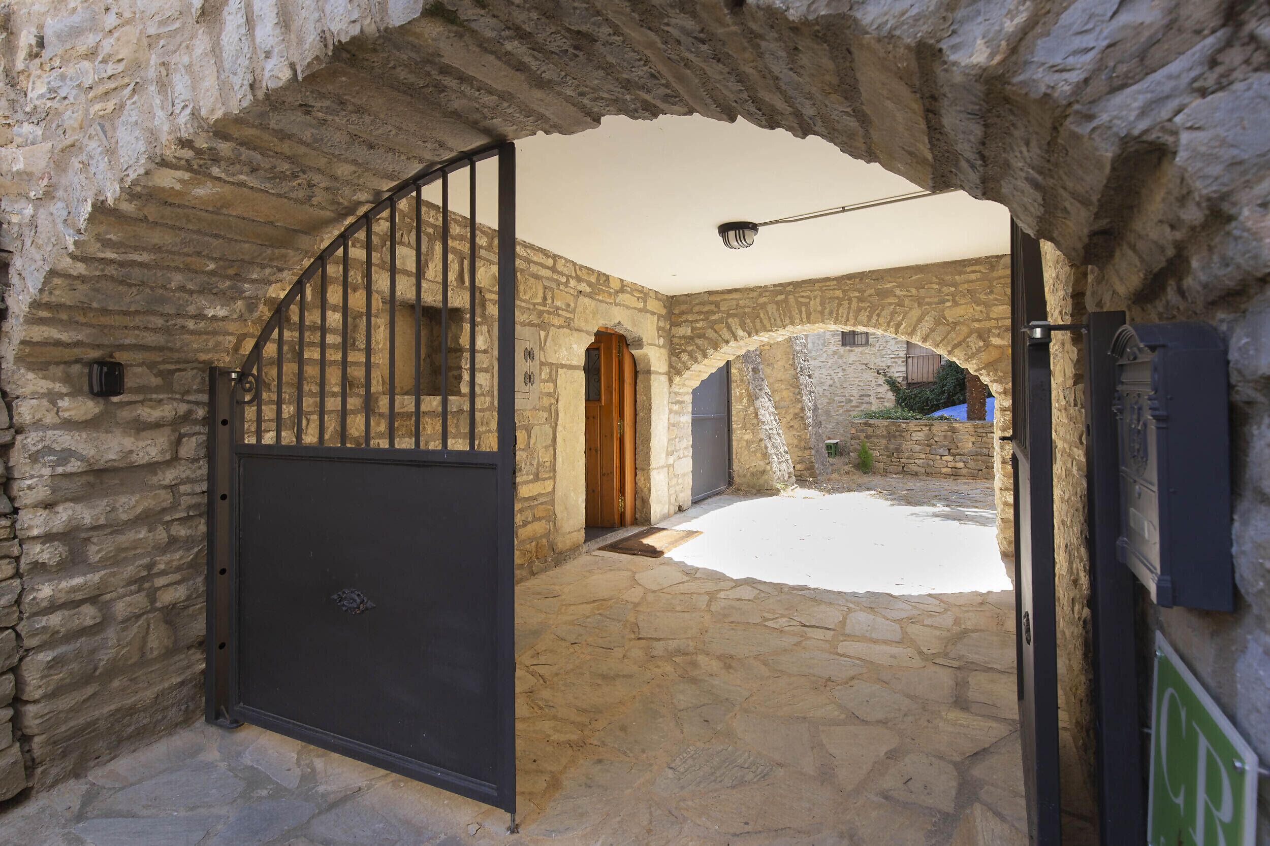 Un portail en pierre en arc de cercle mène à une cour intérieure dotée d’une grille en métal, de murs en pierre et d’une petite porte en bois.