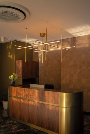 A reception desk with a wooden finish and a metallic accent, a modern pendant light fixture, and a patterned wall in the background.