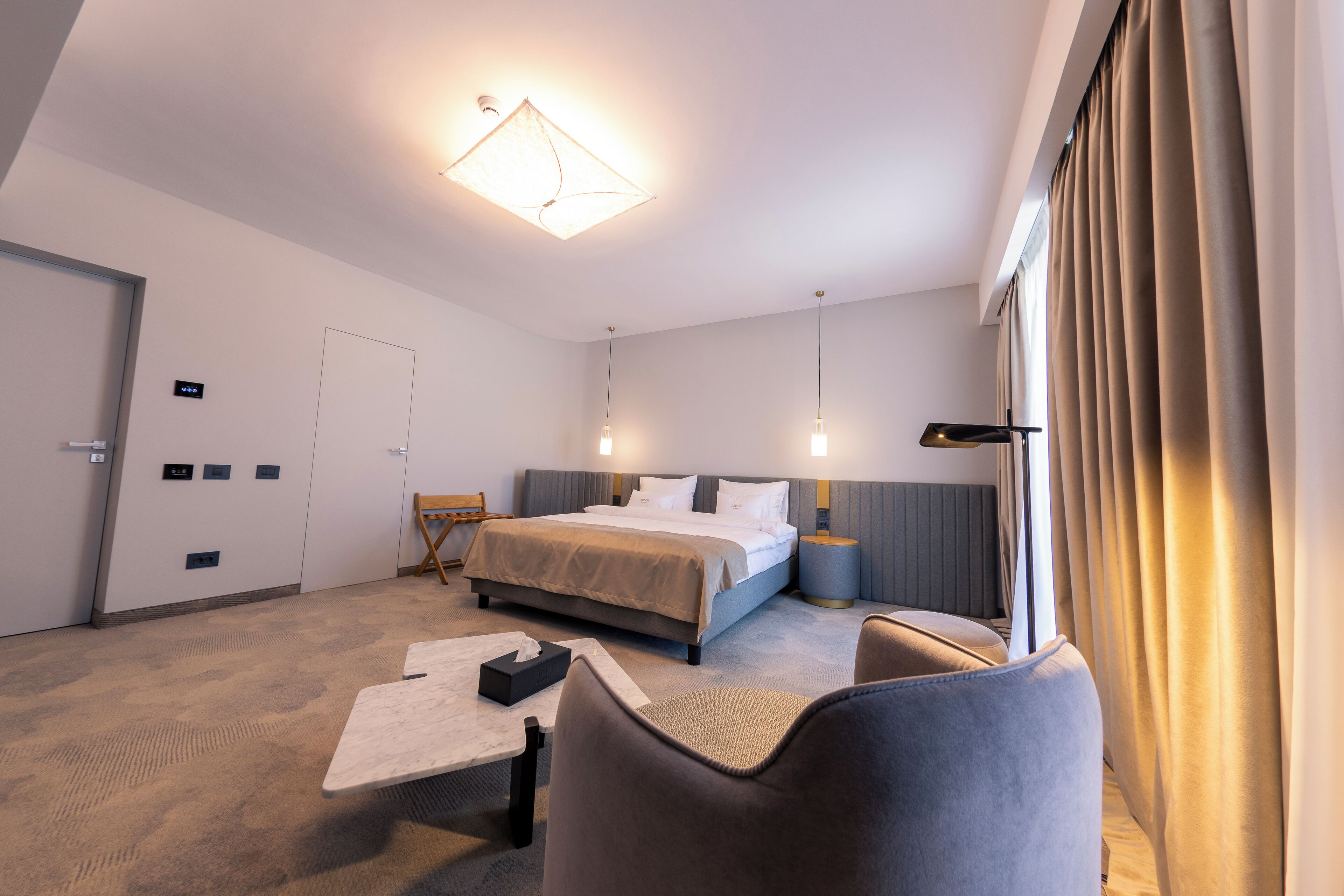 Habitación de hotel con cama, mesitas de noche, escritorio, silla y un amplio ventanal con cortinas.