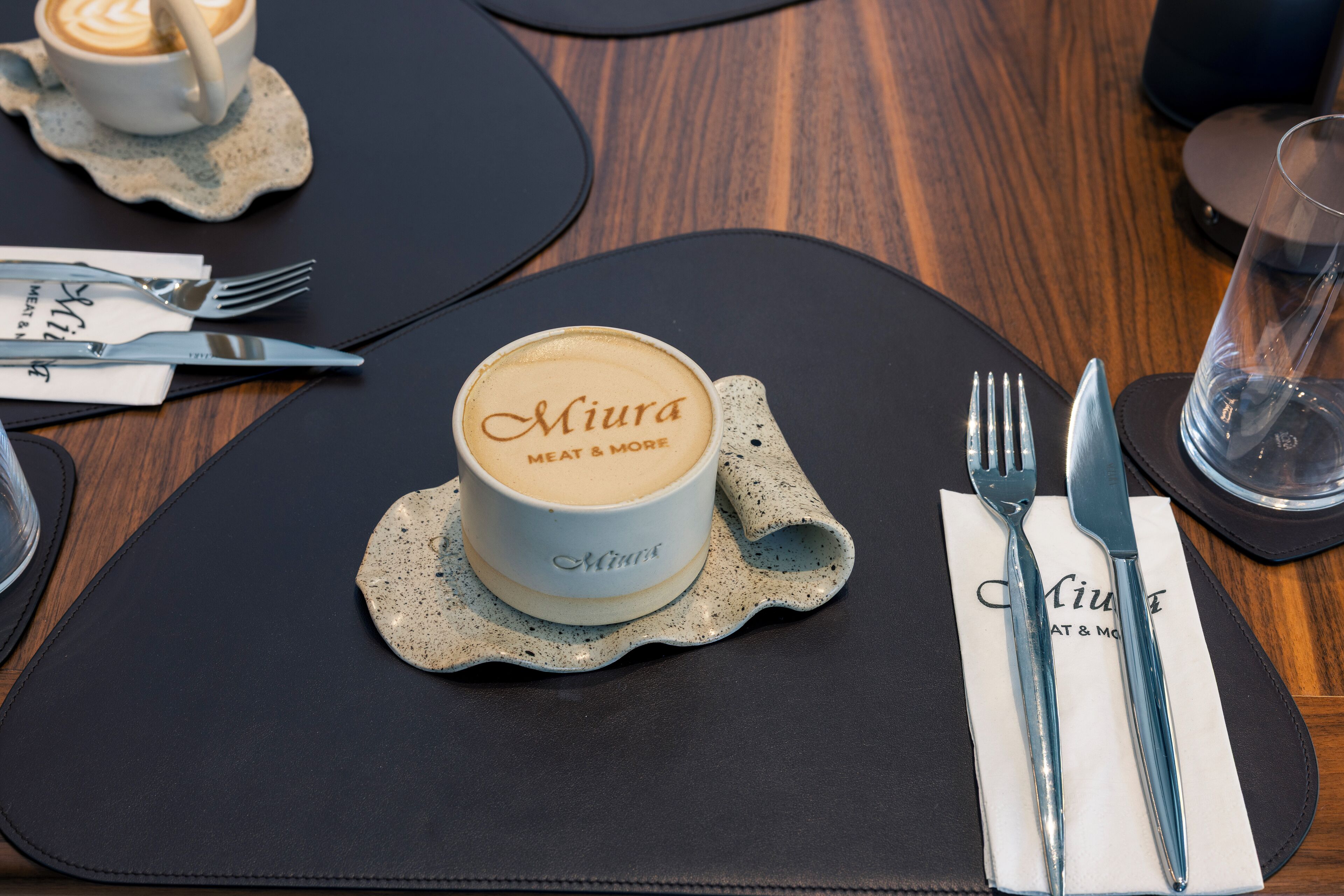 Une table dressée avec une tasse à café portant l’inscription « Miura Meat & More », des couverts et un verre d’eau.