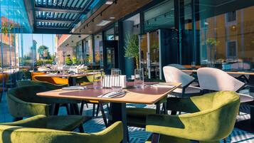 An outdoor dining area with tables and chairs, large windows, and a view of the city.