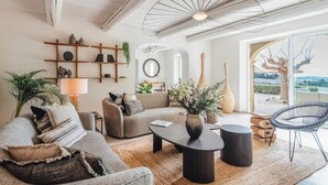 Een moderne woonkamer met een bank, fauteuils, een salontafel en een stoel. Er is een plant, een lamp en een uitzicht op de buitenlucht door een groot raam.