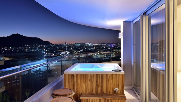 Un balcón moderno con jacuzzi, taburetes de madera y vista a la ciudad al anochecer.