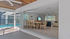Un salón moderno con techo de madera, comedor con sillas de madera, televisor y piscina visible a través de puertas de cristal.