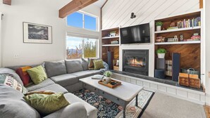 Una sala de estar moderna con un sofá seccional gris, una mesa de centro y una chimenea. Hay estanterías y un televisor montado en la pared.