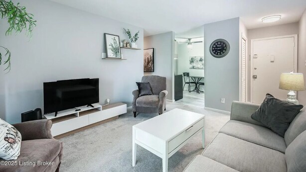 Un salon moderne avec un téléviseur à écran plat, un canapé gris, une table basse blanche et un fauteuil gris. Des tableaux encadrés ornent le mur, et une plante est suspendue au plafond.