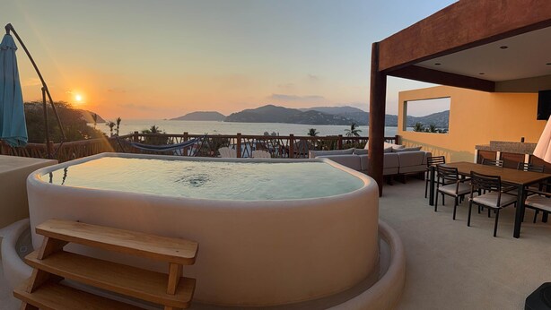Un bain à remous avec un banc en bois, un coin repas avec une table et des chaises, et une vue sur la mer et les montagnes au coucher du soleil.