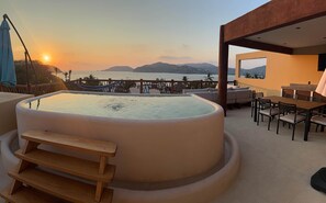 A hot tub with a wooden bench, a dining area with a table and chairs, and a view of the sea and mountains at sunset. - Penthouse private pool 1 block from beach 3 bedroom (Zihuatanejo)