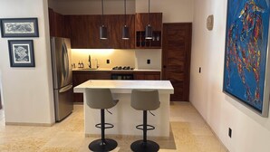 Une cuisine moderne avec des armoires en bois, un réfrigérateur en acier inoxydable, un comptoir blanc, deux tabourets de bar, des luminaires suspendus et un grand tableau abstrait accroché au mur.