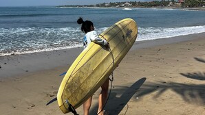 Une personne transportant une planche de surf jaune sur une plage de sable, avec des vagues de l’océan en arrière-plan.