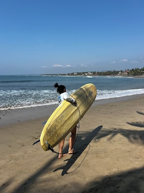 Eine Person trägt einen gelben Surfbrett am sandigen Strand, im Hintergrund sind Meereswellen zu sehen.