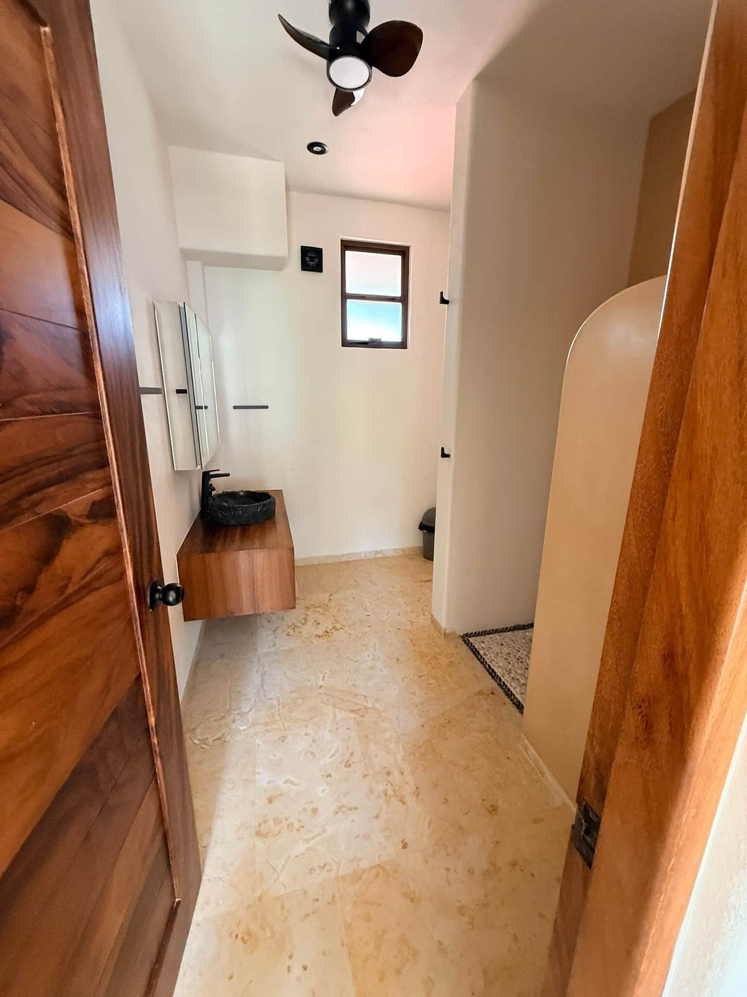 Un pasillo con ventilador de techo, una ventana pequeña y una puerta con marco de madera.
