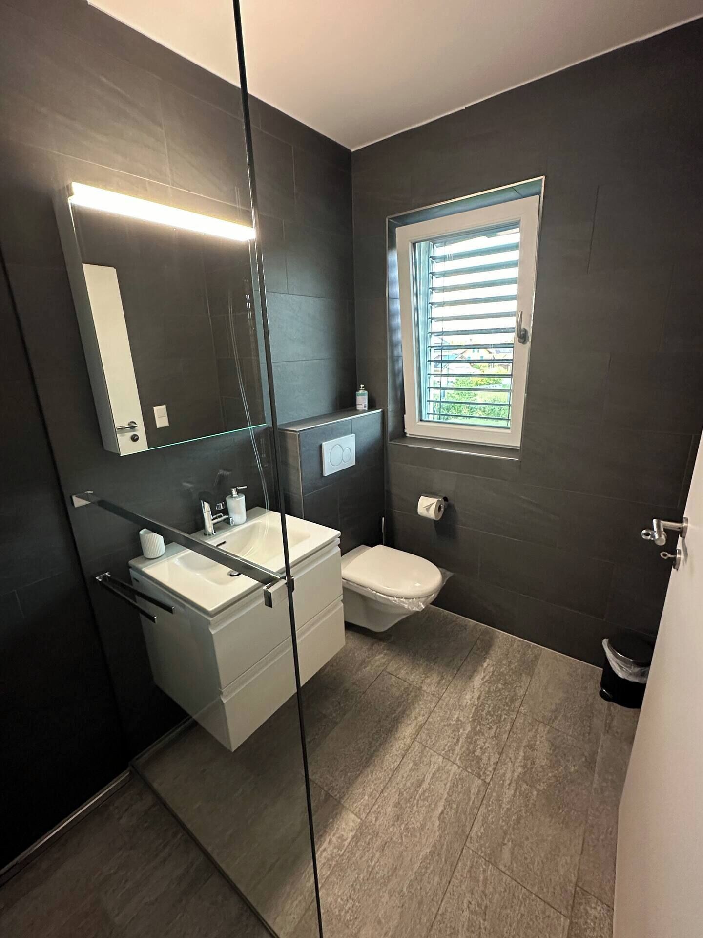 A modern bathroom with a glass shower enclosure, a white vanity with a mirror, a toilet, and a window with blinds.
