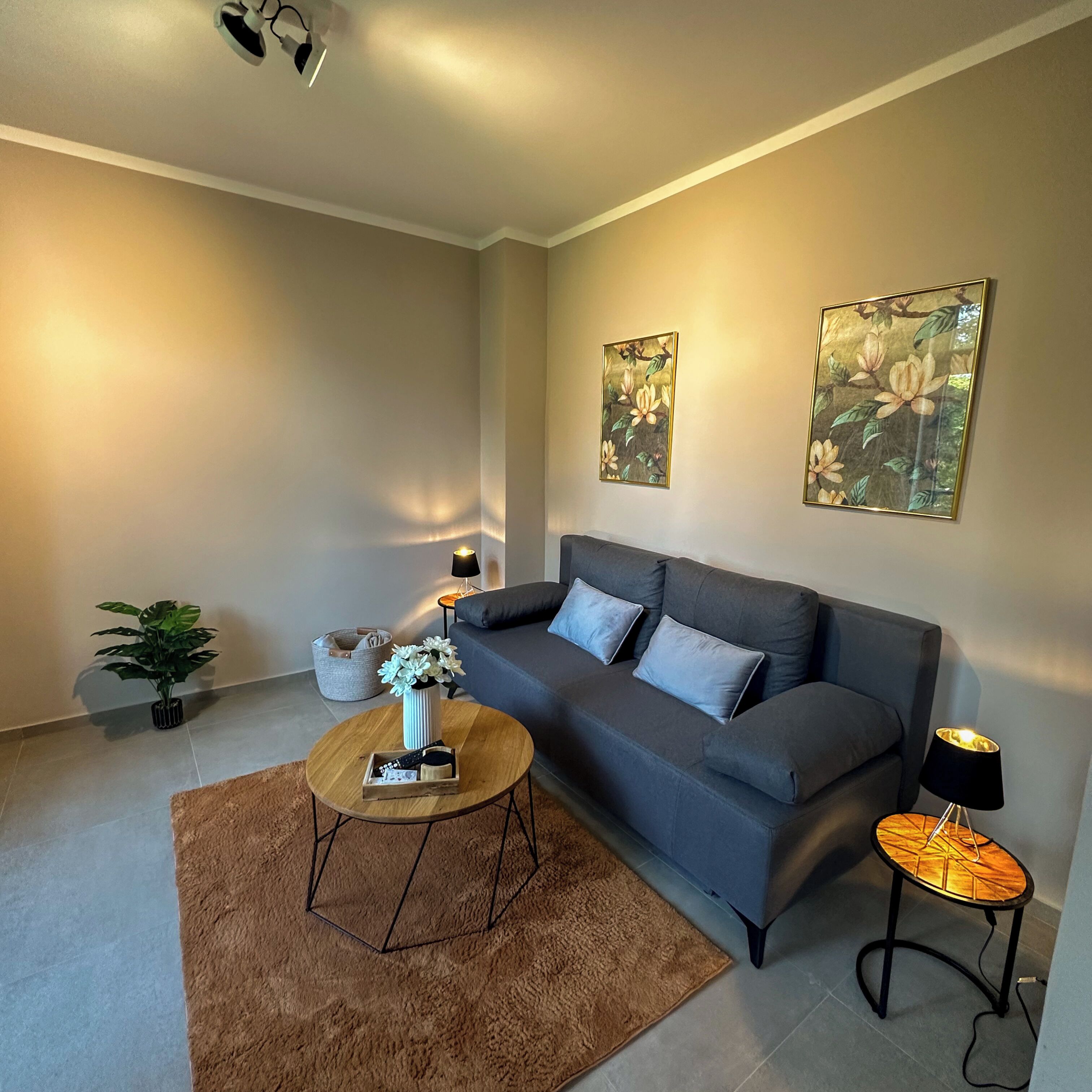Un salon moderne avec un canapé gris, une table basse ronde, une petite table d’appoint et deux tableaux accrochés au mur.