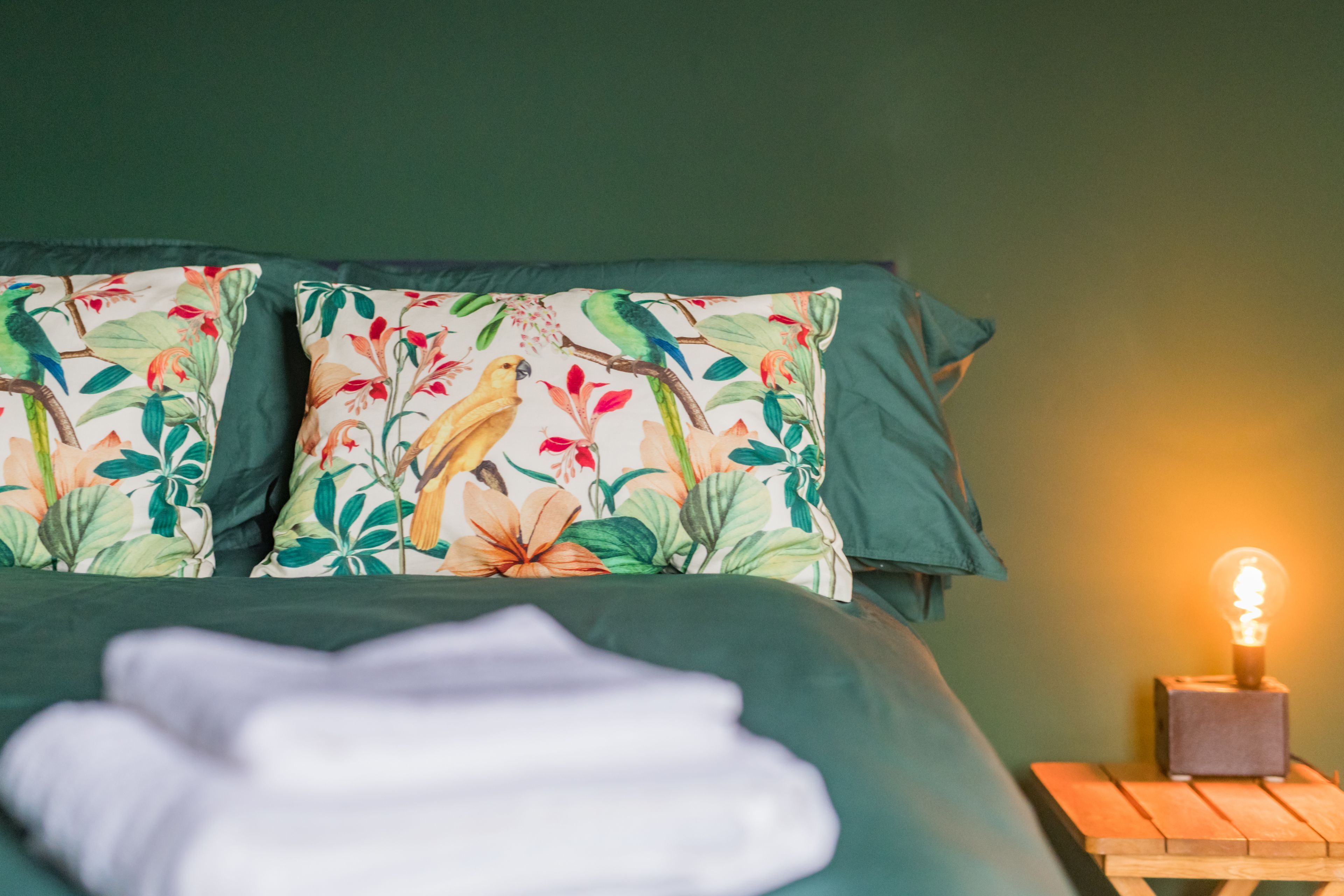 A bed with floral pillows, a bedside table with a light bulb, and a green wall.