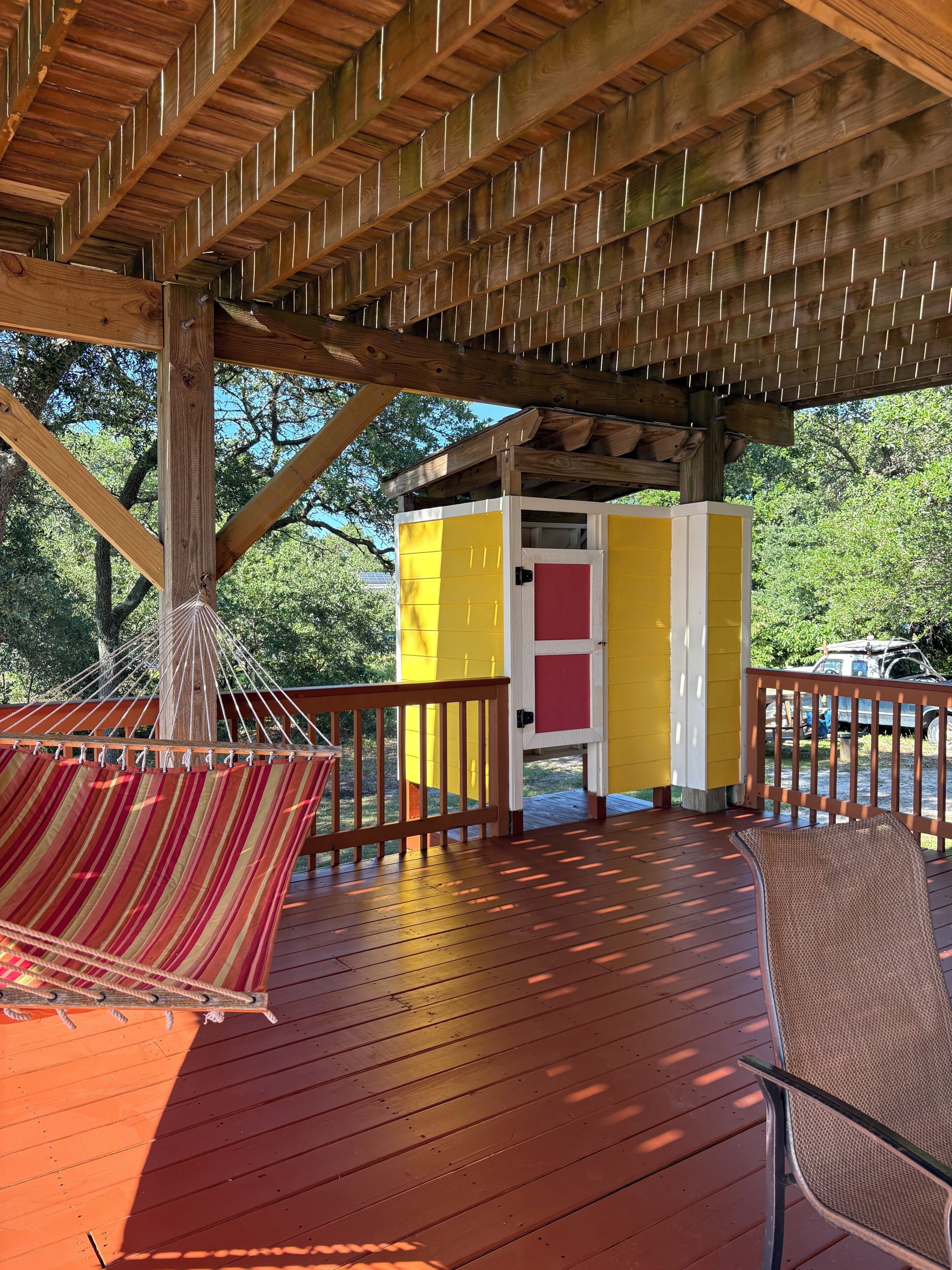 Une terrasse en bois avec un hamac, un bâtiment jaune et rouge, et un véhicule en arrière-plan.