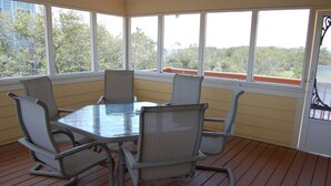 A covered patio with a glass table and four chairs, large windows offering a view of trees and buildings.