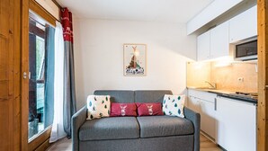 A cozy living area with a grey sofa, a poster on the wall, and a kitchen with white cabinets and a microwave. - Le Cocon des Pistes - Station à 10 minutes (Les Contamines-Montjoie)