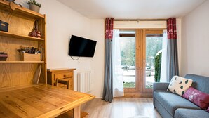 A cozy living room with a wooden dining table, a grey sofa with decorative pillows, a flat-screen TV mounted on the wall, and a window with curtains. - Le Cocon des Pistes - Station à 10 minutes (Les Contamines-Montjoie)