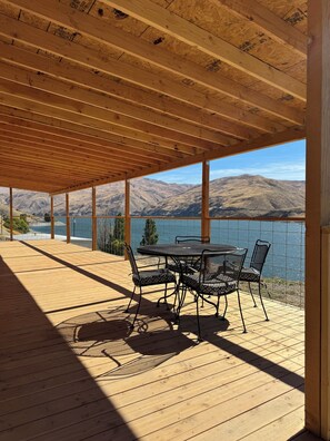 Une terrasse extérieure couverte, dotée d’une table et de chaises, qui offre une vue sur un plan d’eau et des montagnes.
