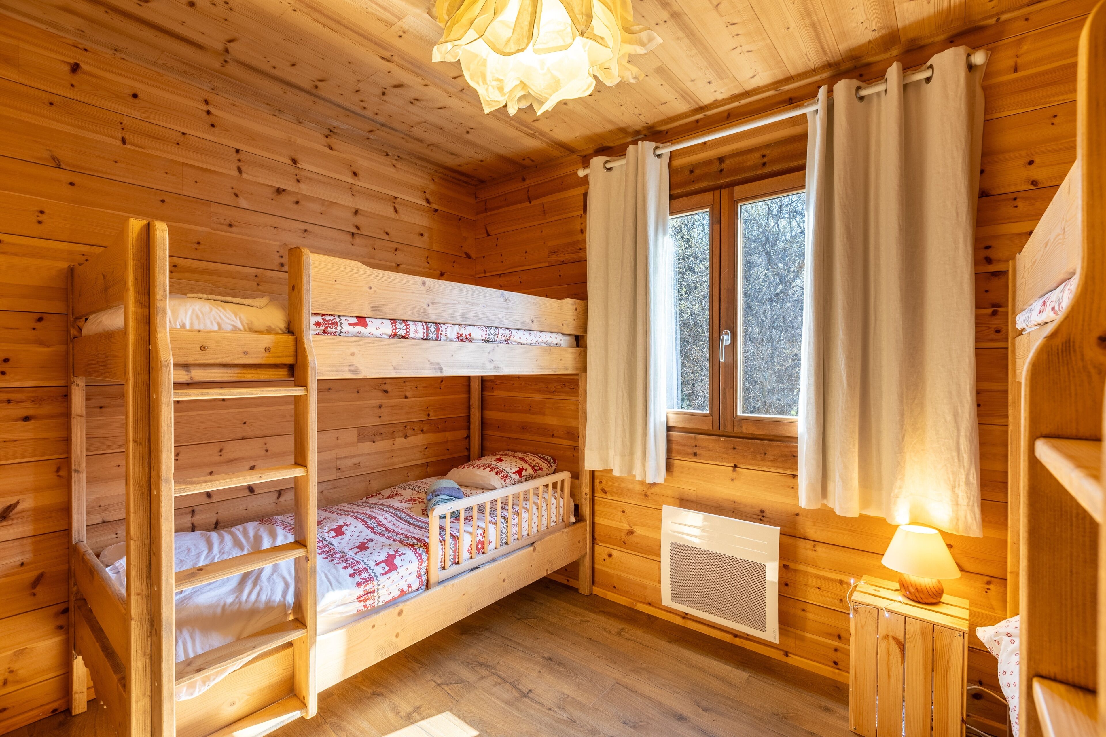 A wooden cabin room with bunk beds, a window with curtains, and a small bedside table with a lamp.