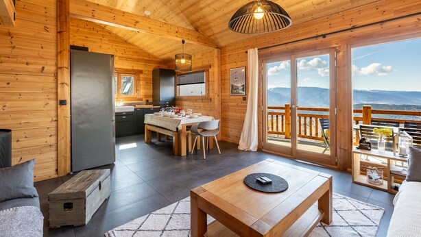 En stuga med träpanel på insidan, innehållande en matplats, ett kök och en balkong med utsikt över bergen.